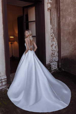 The bride, in the elegant River gown by Modeca, featuring a flowing white design with an intricate back, stands gracefully in a doorway adorned with ornate stonework, capturing the essence of timeless elegance.