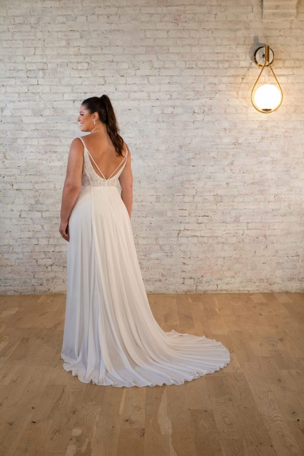 A woman in a Stella York 7579 - Pearla gown stands on the wooden floor, facing a brick wall with a hanging light.