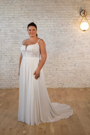 A woman in a Stella York 7579 - Pearla white wedding dress stands gracefully in front of a rustic brick wall, illuminated by a vintage hanging light fixture.