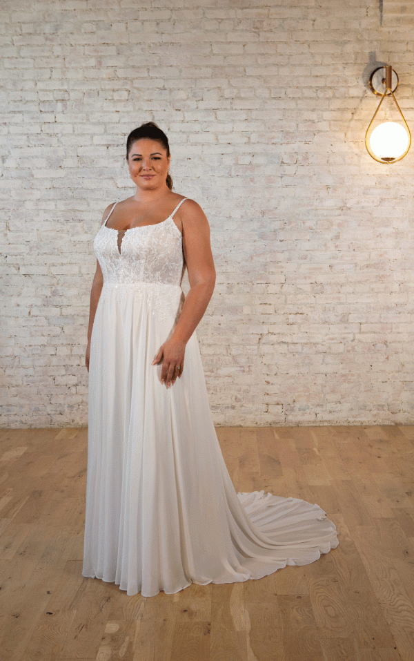 A woman in a Stella York 7579 - Pearla white wedding dress stands gracefully in front of a rustic brick wall, illuminated by a vintage hanging light fixture.