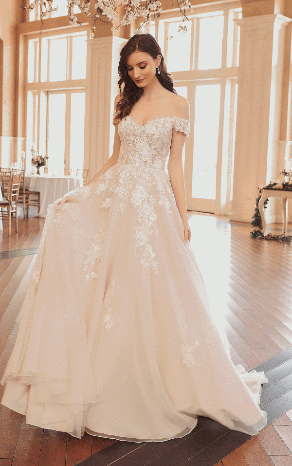 Bride in the elegant Justin Alexander 44312 - Jasmine off-shoulder lace wedding gown poses gracefully in a bright room with large windows and wooden floors, evoking the essence of a jasmine-filled garden.