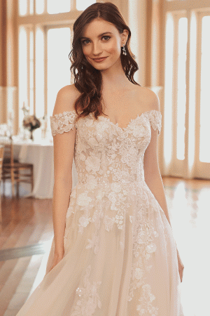 The bride, adorned in a breathtaking Justin Alexander 44312 - Jasmine lace wedding dress, stands gracefully in a bright room with wooden floors and expansive windows.