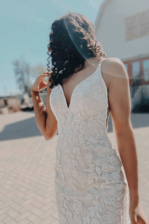 In the outdoor sunlight, a bride stands in a stunning Stella York 7312 - April bridal gown with her back partially turned, highlighting the intricate lace detailing.