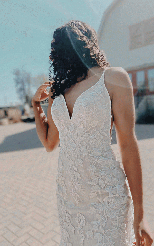 In the outdoor sunlight, a bride stands in a stunning Stella York 7312 - April bridal gown with her back partially turned, highlighting the intricate lace detailing.