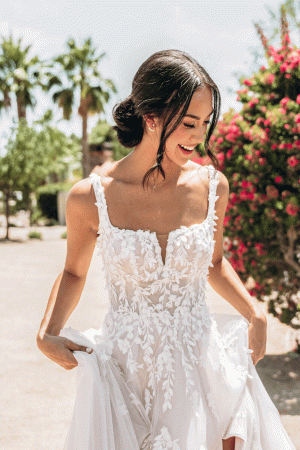 A bride in the Stella York 7965 - Tiger Lilly floral lace wedding gown smiles outdoors amid vibrant tiger lilies, trees, and other colorful blooms.