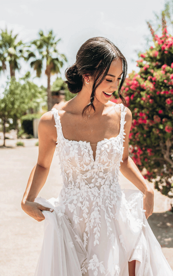 A bride in the Stella York 7965 - Tiger Lilly floral lace wedding gown smiles outdoors amid vibrant tiger lilies, trees, and other colorful blooms.