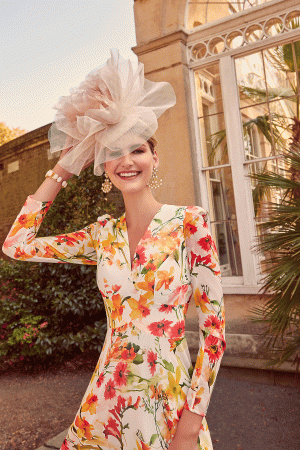 A woman in a floral dress and large hat stands smiling outdoors, her vivid attire echoing the joyful vibes of Invitations by Veni - 29803, near a building with striking glass windows.