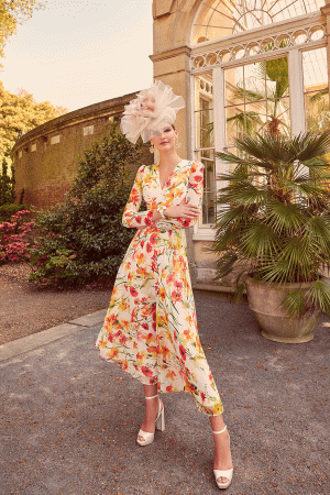 Draped in a floral dress from Invitations by Veni - 29803 and wearing a large hat, the woman stands gracefully in the garden, her heels gently sinking into the lush grass. An air of elegance envelops her while an old stone building rises behind, its presence reminiscent of a bygone era's silent call.