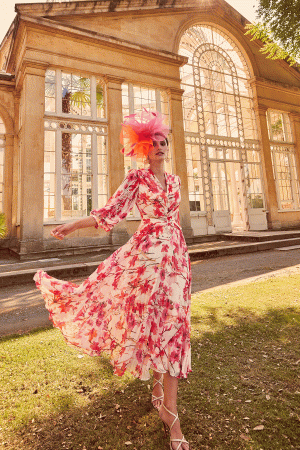 A woman in a floral dress and pink hat poses elegantly in front of a glass conservatory, as if she awaits her moment worthy of Invitations by Veni - 29803.