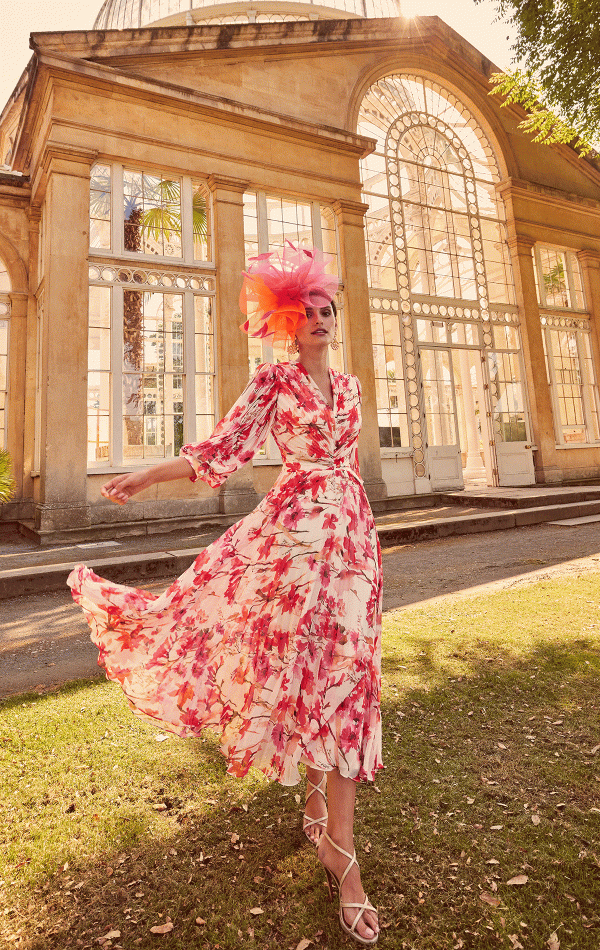 A woman in a floral dress and pink hat poses elegantly in front of a glass conservatory, as if she awaits her moment worthy of Invitations by Veni - 29803.