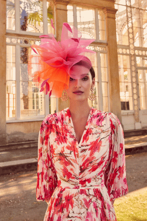 A woman wearing a floral dress and a large pink hat stands gracefully in front of a glass building, emanating elegance like the allure of "Invitations by Veni - 29803," inviting you into her world.