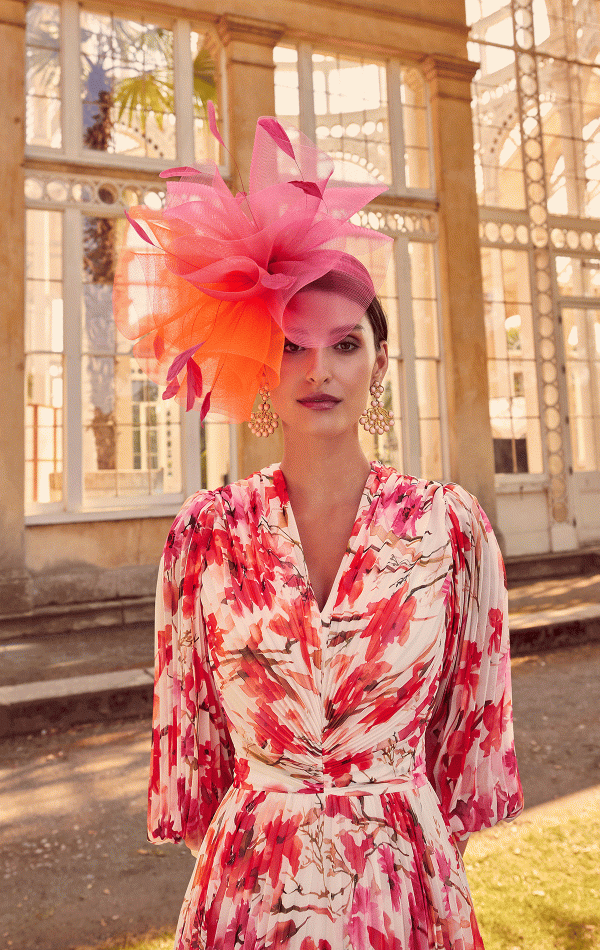 A woman wearing a floral dress and a large pink hat stands gracefully in front of a glass building, emanating elegance like the allure of "Invitations by Veni - 29803," inviting you into her world.