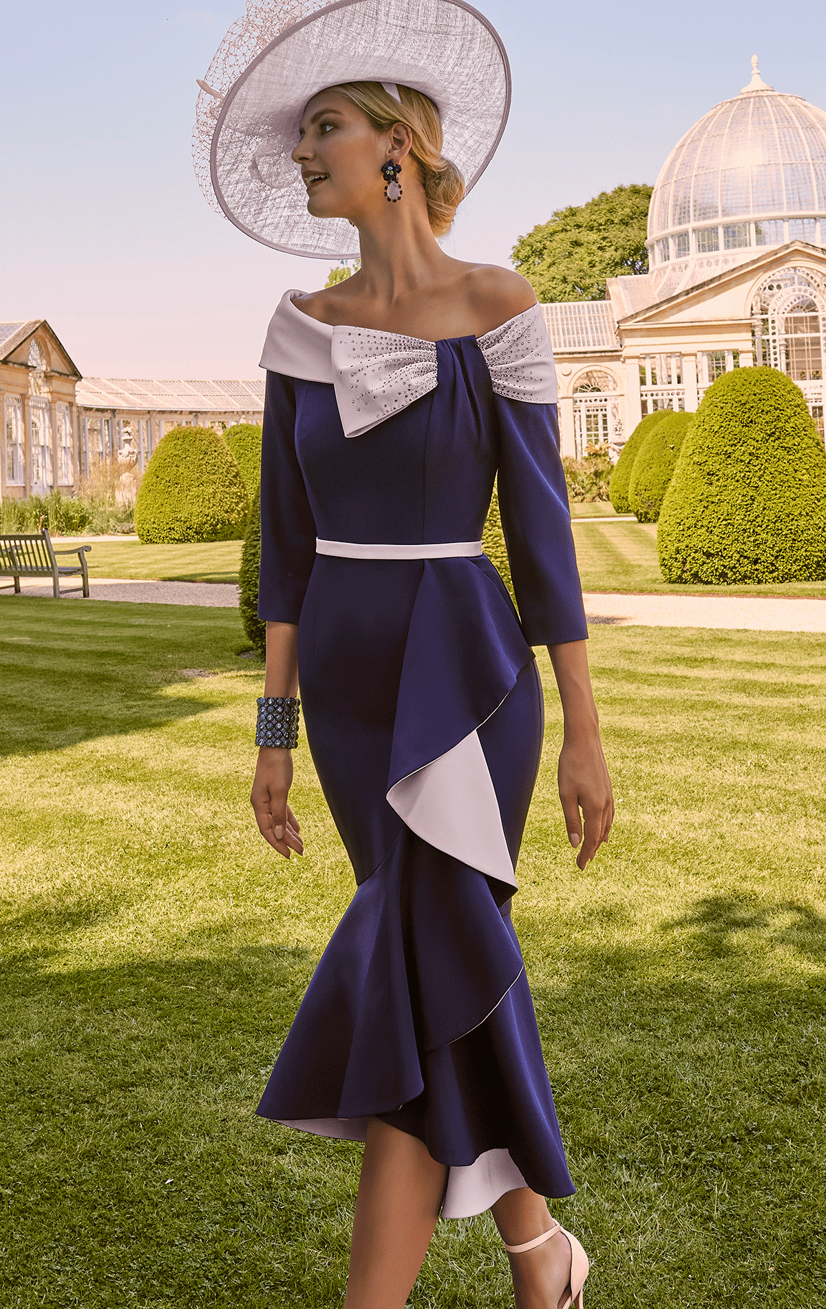 A woman in an elegant blue and white dress and hat gracefully strolls through a garden near a glass conservatory, as if beckoned by the silent allure of nature’s Invitations by Veni - 29803.