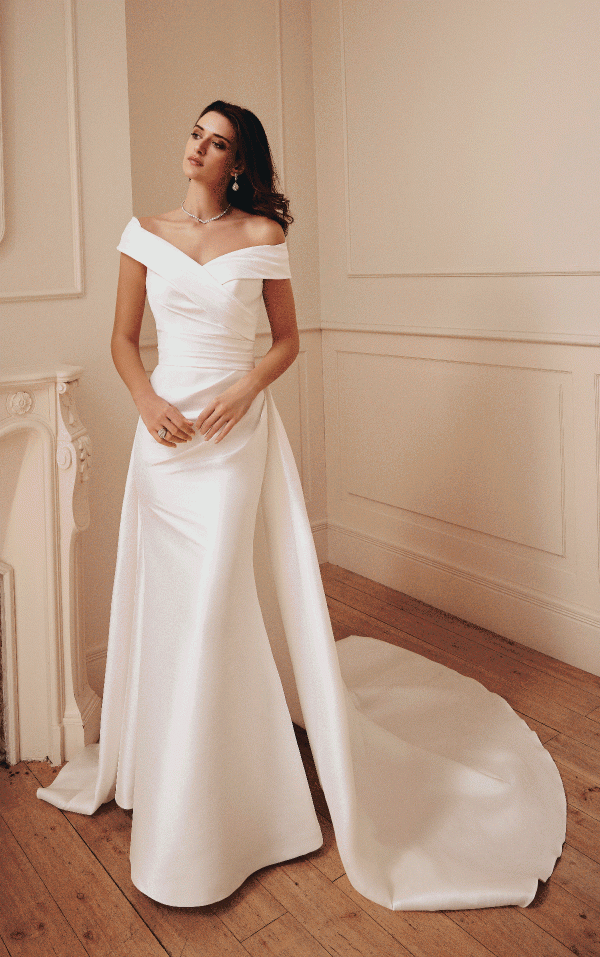 In a softly lit room, a woman stands gracefully in an elegant Victoria Jane 18744 - Germain satin off-shoulder wedding gown, complete with a long train.