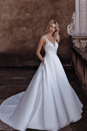 A woman in a white Modeca River wedding dress stands in front of an ornate stone wall, with the faint sound of a river flowing gently nearby.