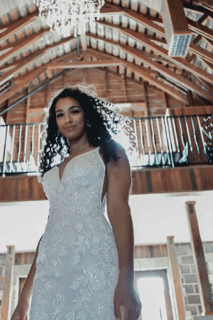 Wearing a stunning Stella York 7312 - April bridal gown, the bride stands elegantly in a rustic venue, surrounded by wooden beams and illuminated by a chandelier above.