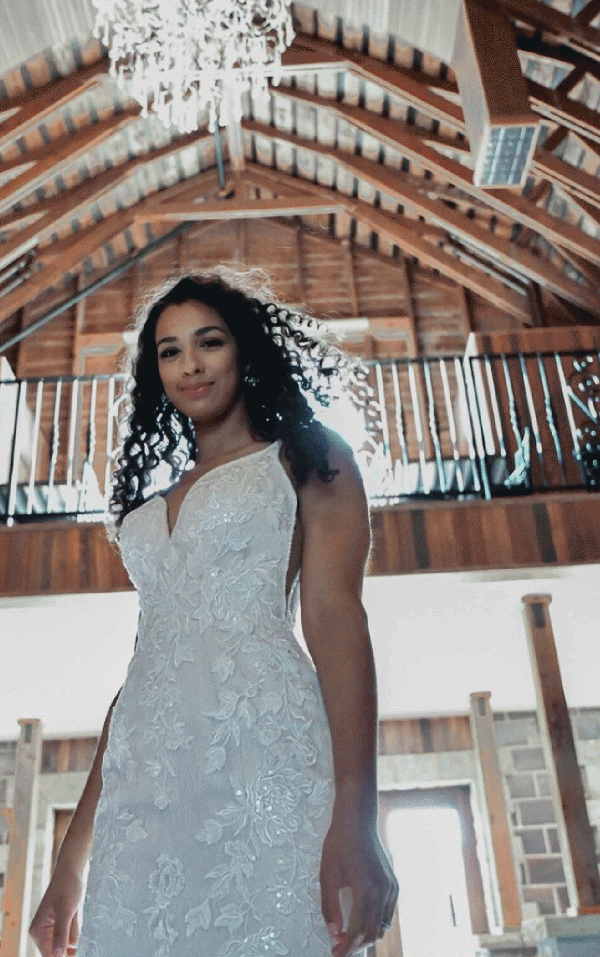 Wearing a stunning Stella York 7312 - April bridal gown, the bride stands elegantly in a rustic venue, surrounded by wooden beams and illuminated by a chandelier above.