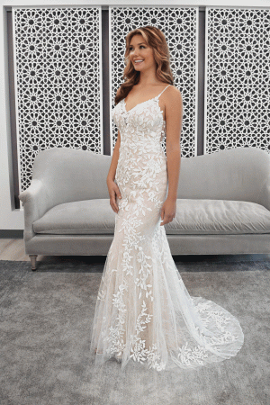 A smiling woman in a Stella York 7312 - April bridal gown stands gracefully in front of a decorative wall, with a gray couch nearby.