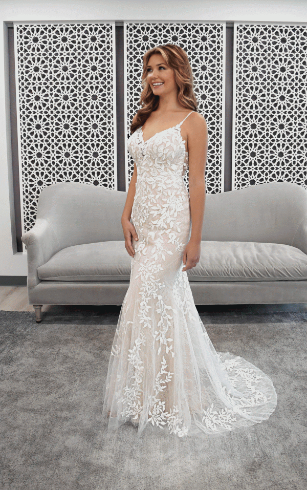 A smiling woman in a Stella York 7312 - April bridal gown stands gracefully in front of a decorative wall, with a gray couch nearby.