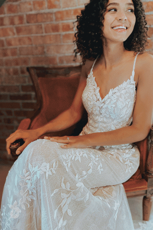 A woman in a stunning Stella York 7312 - April bridal gown sits on a chair, smiling against a brick wall background, capturing the essence of an April wedding.