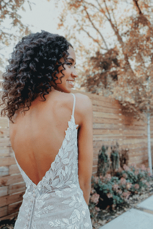 In the gentle embrace of April, a woman stands gracefully in a delicate lace dress with an open back. The wooden fencing and trees frame her elegance, echoing the exquisite design of the Stella York 7312 - April.