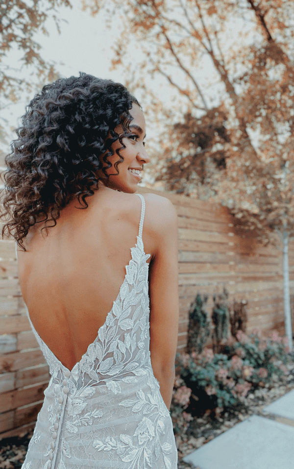 In the gentle embrace of April, a woman stands gracefully in a delicate lace dress with an open back. The wooden fencing and trees frame her elegance, echoing the exquisite design of the Stella York 7312 - April.