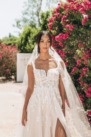 The bride, adorned in the exquisite Stella York 7965 - Tiger Lilly white lace dress and veil, stands gracefully amidst vibrant pink flowers outdoors, embodying timeless elegance.