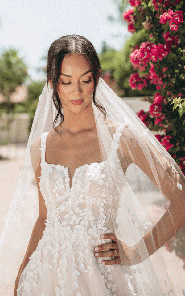 Wearing the Stella York 7965 - Tiger Lilly wedding dress and a veil, the bride stands outdoors, surrounded by a vibrant array of Tiger Lilly flowers.