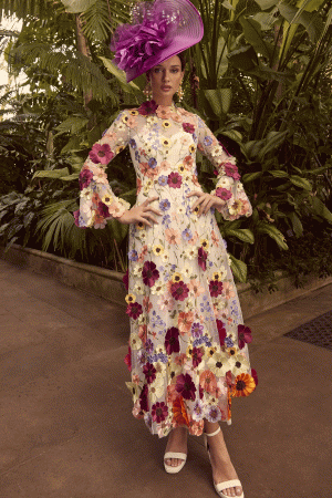 A woman wearing the Veni Infantino 992345 dress and a purple hat poses gracefully amid a lush garden setting.