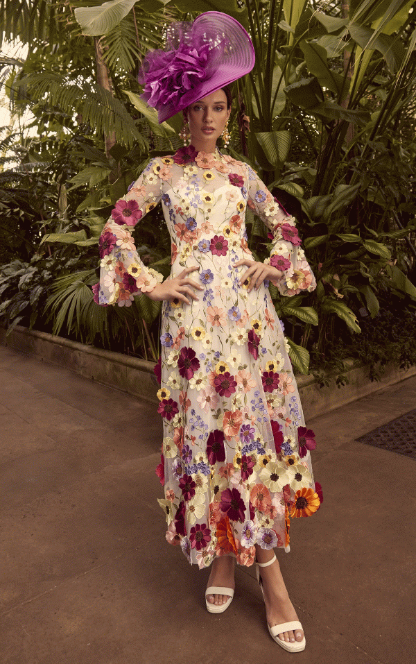 A woman wearing the Veni Infantino 992345 dress and a purple hat poses gracefully amid a lush garden setting.