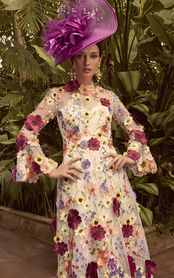 In a lush garden setting, a person wearing the Veni Infantino - 992345 floral dress and a large purple hat poses confidently, exuding elegance.