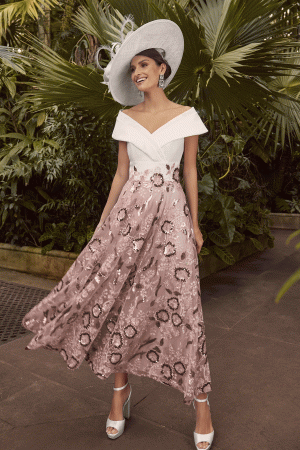 Dressed in the sophisticated Veni Infantino - 992351 ensemble, a woman in a white off-shoulder top paired with a pink floral skirt stands gracefully amid a lush garden, her wide-brimmed hat lending an air of elegance.