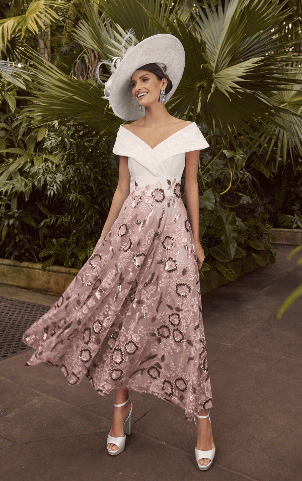 Dressed in the sophisticated Veni Infantino - 992351 ensemble, a woman in a white off-shoulder top paired with a pink floral skirt stands gracefully amid a lush garden, her wide-brimmed hat lending an air of elegance.