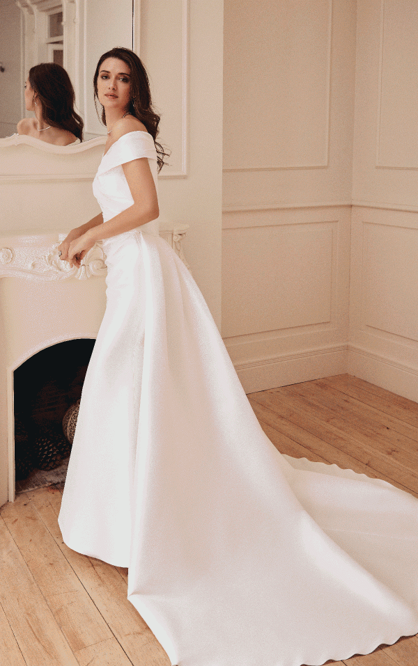 A woman in the sophisticated Victoria Jane 18744 - Germain off-shoulder wedding dress poses by a fireplace in a room with light-colored walls and a wooden floor, exuding timeless charm.