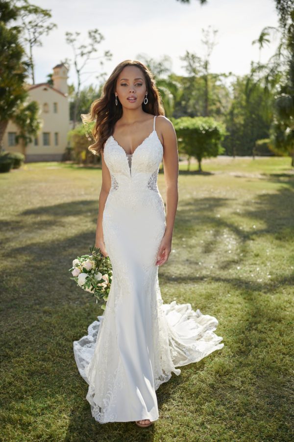 A bride in a stunning Stella York 7118 - Sky wedding dress holds flowers while walking on the grass beneath a clear sky, with lush trees and a charming house in the background.