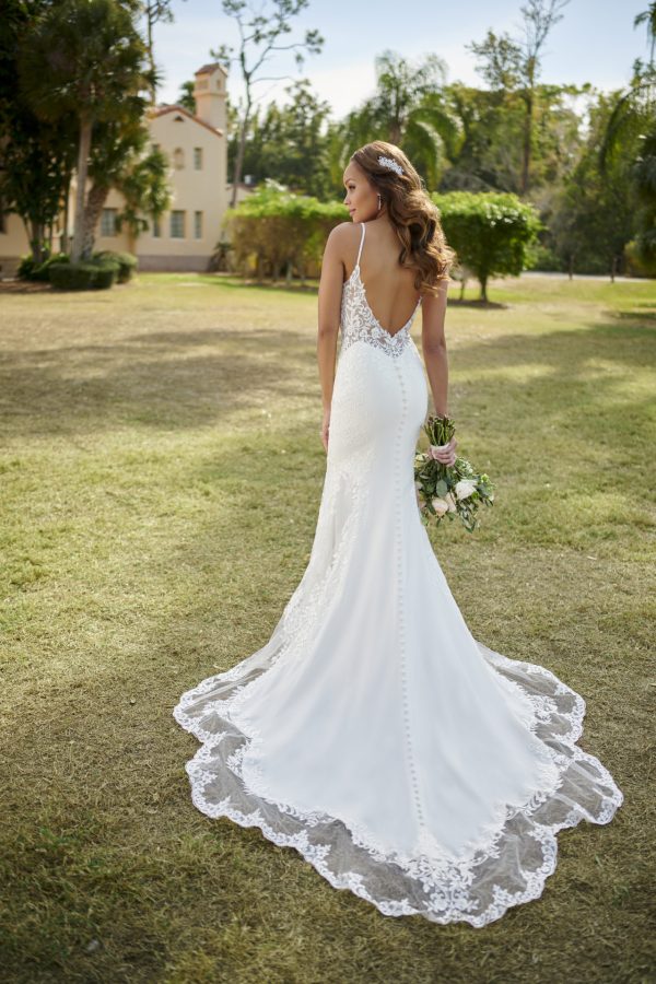 The bride, in a stunning Stella York 7118 - Sky lace wedding gown, holds her bouquet gracefully while standing on lush grass. With trees and a building in the background, the scene is beautifully completed by a clear blue sky.