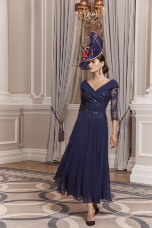 A woman in an elegant blue Veni Infantino - 992013 dress with a broad hat stands gracefully in a sophisticated room adorned with ornate curtains.