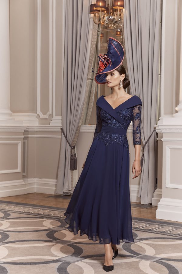 A woman in an elegant blue Veni Infantino - 992013 dress with a broad hat stands gracefully in a sophisticated room adorned with ornate curtains.