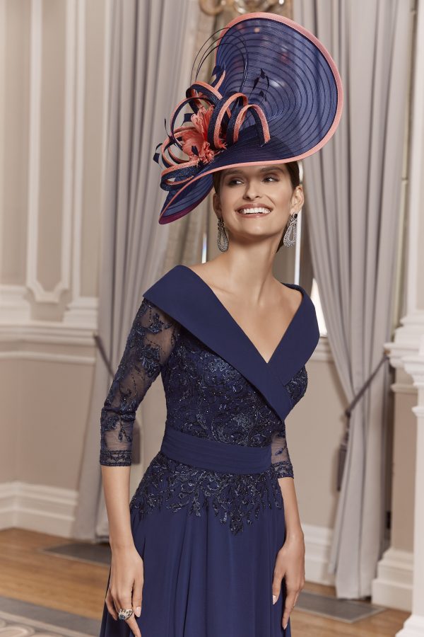 A woman in an elegant navy dress from the Veni Infantino collection, style 992013, paired with a large hat, smiles warmly in a room adorned with draped curtains.