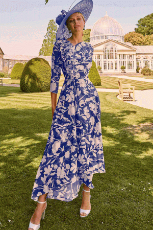 Wearing a blue floral dress and hat from Invitations by Veni - 36069, she stands gracefully on the grass with an elegant garden scene and a charming greenhouse in the background.