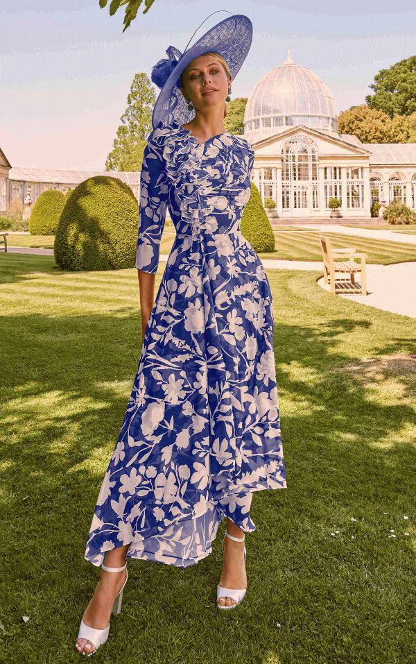 Wearing a blue floral dress and hat from Invitations by Veni - 36069, she stands gracefully on the grass with an elegant garden scene and a charming greenhouse in the background.