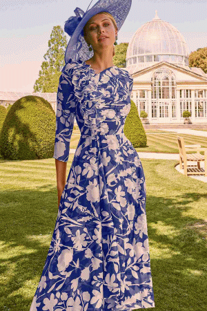 A woman wearing a blue and white floral dress with a matching hat stands in a garden, holding "Invitations by Veni - 36069," with a glasshouse visible in the background.