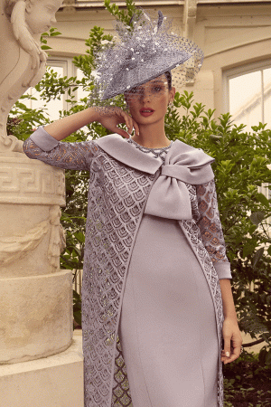 In a sunlit conservatory, a woman in an elegant lilac Veni Infantino 992314 dress and hat poses gracefully near a statue and lush greenery.