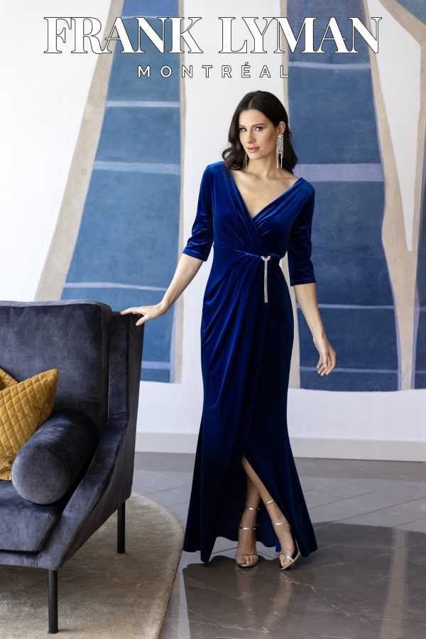 A woman elegantly stands by a modern couch, wearing a stunning Frank Lyman 239236 blue velvet dress, with geometric wall art in the background.