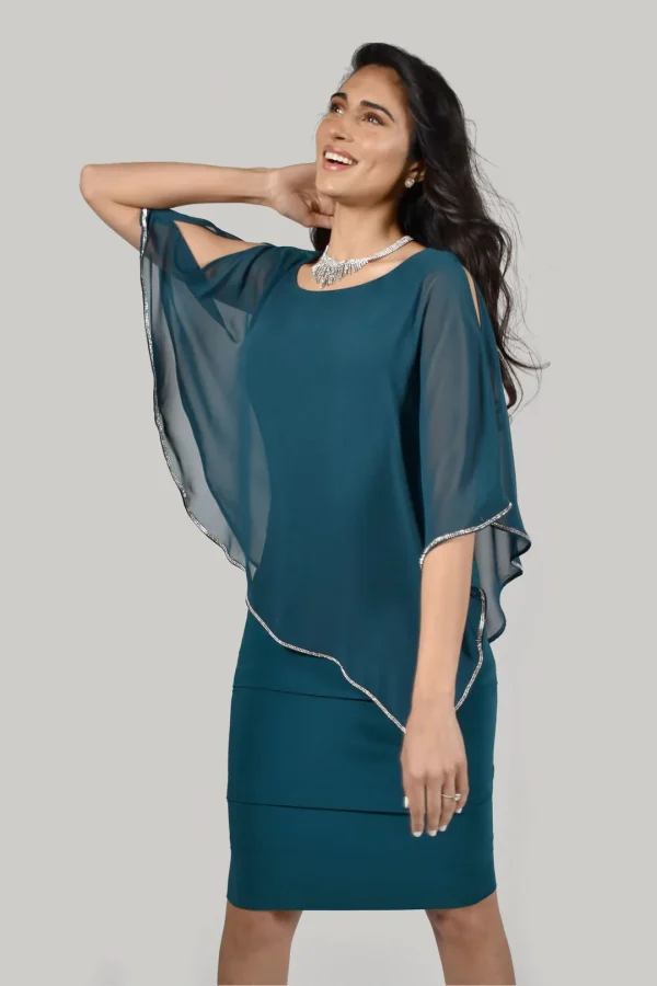 A woman in a stylish teal dress with a sheer overlay, featuring the stunning Frank Lyman - 219022 design, smiles radiantly while posing with her hand behind her head, embodying effortless fashion elegance.