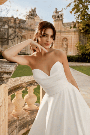 Inma, wedding dress 44495, poses outdoors in a white strapless gown, elegantly set against a stone railing with historic architecture as the stunning backdrop.