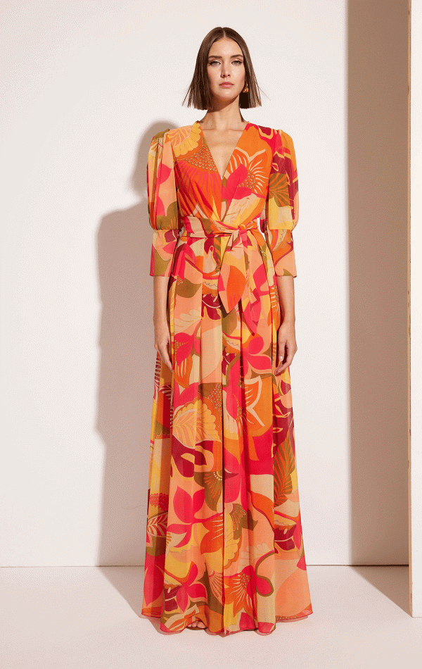 In a vibrant Carla Ruiz - 51630 floral dress, a woman stands poised against a plain background, hands at her sides as she gazes forward.