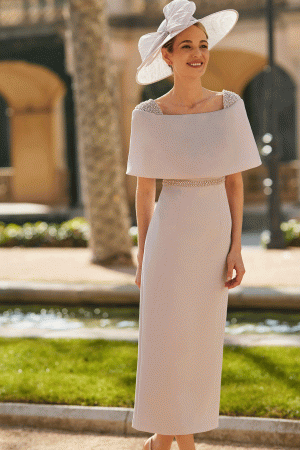 A woman in an elegant white Rosa Clara Couture Club - 9G99 dress and wide-brim hat smiles outdoors, surrounded by lush greenery and stunning architecture.