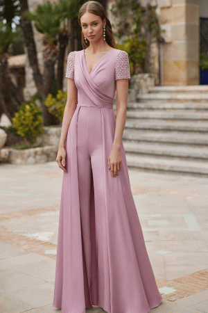 A woman in a pink Rosa Clara Couture Club 9G99 jumpsuit with embellished sleeves stands outdoors near steps and lush greenery.