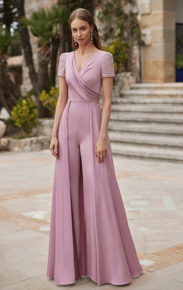 A woman in a pink Rosa Clara Couture Club 9G99 jumpsuit with embellished sleeves stands outdoors near steps and lush greenery.